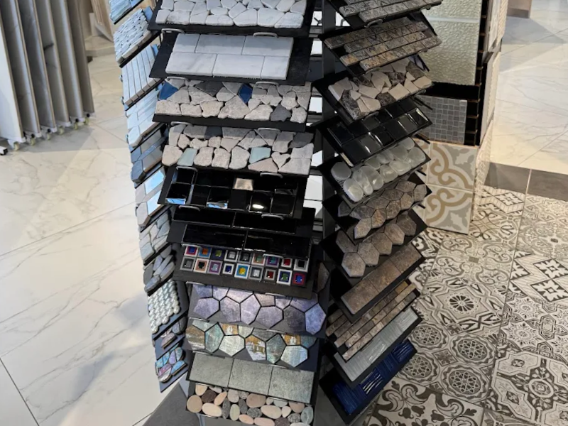 Display of stacked tile samples in varying colors and patterns, arranged in a tall rack with geometric and mosaic pieces, on a light marble floor with decorative rug.